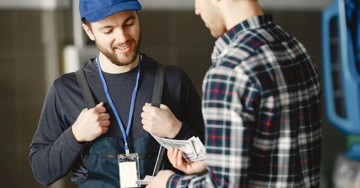Un mécanicien en casquette bleue reçoit un paiement en espèces d'un client en chemise à carreaux, souriant.